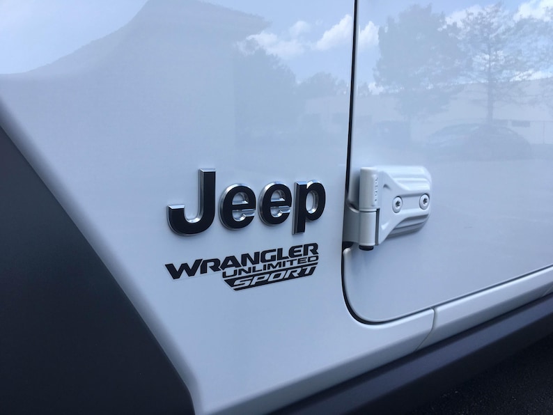 May include: Close-up of a white Jeep Wrangler Unlimited Sport. The Jeep logo is in silver, with the Wrangler Unlimited Sport text below it in black. The door hinge is visible on the right side.