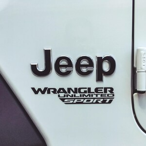 May include: Close-up of a white vehicle panel with the black and chrome Jeep logo and the words "WRANGLER UNLIMITED SPORT" in black lettering. The vehicle's door hinge is visible on the right side.