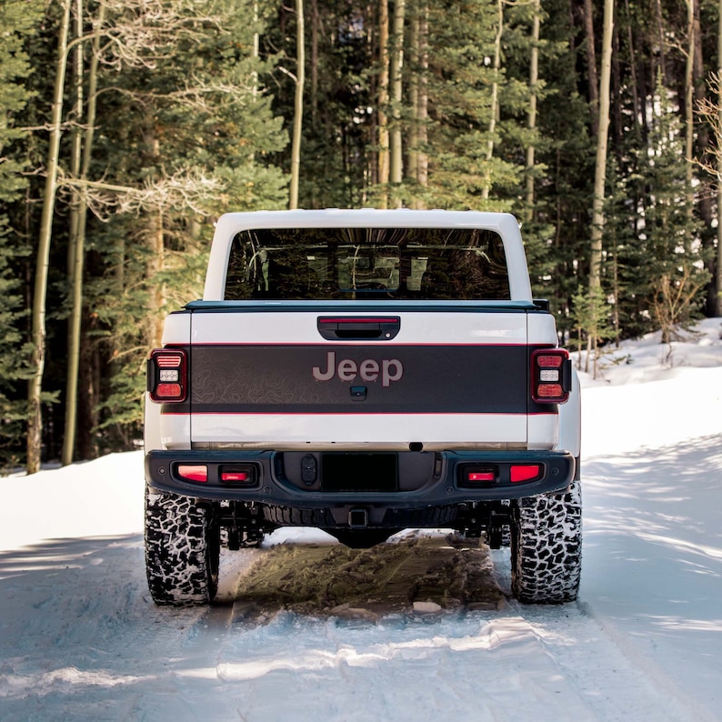 May include: A white Jeep truck with a black and red stripe on the tailgate. The truck is parked in a snowy forest setting.