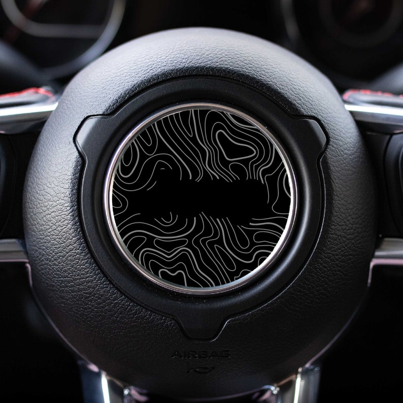 May include: Black leather steering wheel with a silver trim and a black and white topographic design in the center.