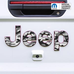 Puede incluir: La palabra "Jeep" en letras de camuflaje grises y blancas con un contorno rosa oscuro. Las letras están adheridas a una superficie blanca. La imagen también incluye un logotipo de Mopar y el texto "OFFICIAL LICENSED PRODUCT".