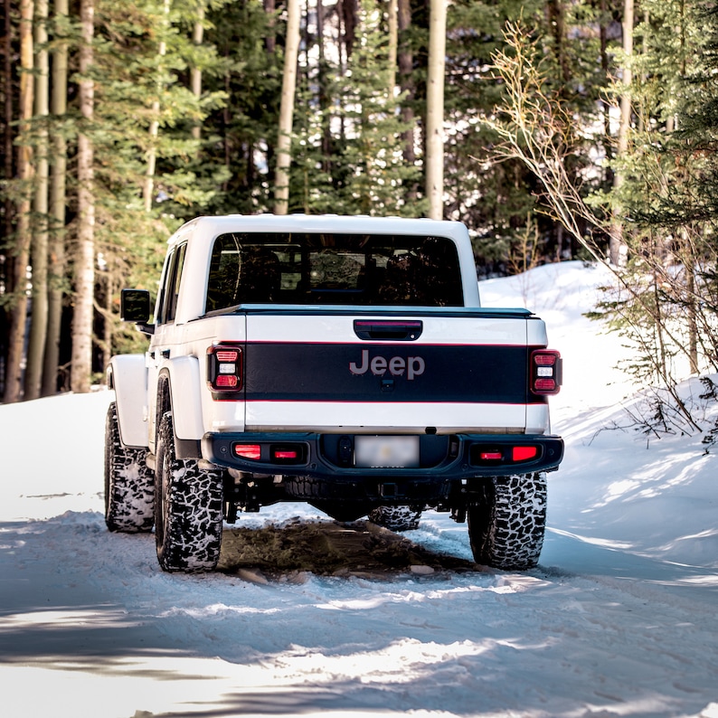 May include: A white Jeep pickup truck with a black tailgate and the word "Jeep" in black lettering on the tailgate. The truck is parked in a snowy forest setting.