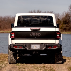 May include: White Jeep pickup truck with a black tailgate and the word "Jeep" in red lettering on the back. The truck is parked on a dirt road with a body of water in the background.