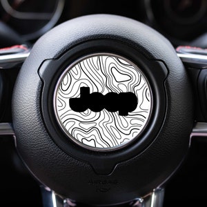 May include: Black and white topographic map design Jeep steering wheel cover. The cover features a black Jeep logo on a white background with black topographic lines.