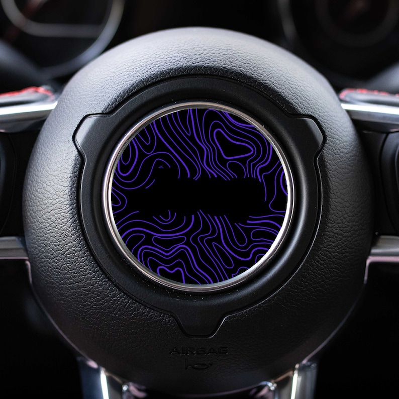 May include: A black car steering wheel with a purple and black patterned car vent cover.