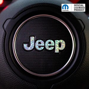 May include: Close-up of a black steering wheel with a silver trim and a textured center. The word "Jeep" is displayed in iridescent, glittery letters. The official Mopar licensed product logo is visible.