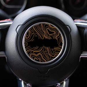 May include: A black car steering wheel with a yellow and black patterned design in the center.