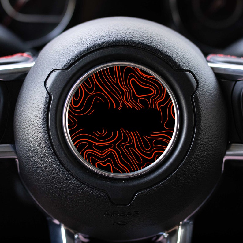 May include: A black car steering wheel with a red and black patterned insert. The insert has a swirling, topographic design.