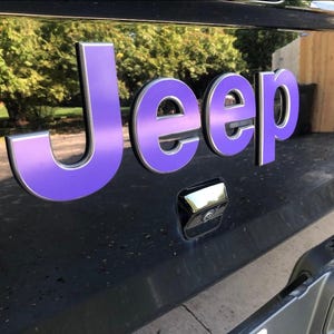 May include: Close-up of a black vehicle's rear with a purple and silver "Jeep" emblem. The emblem is three-dimensional and has a metallic finish. The background shows a blurred view of trees and a building.