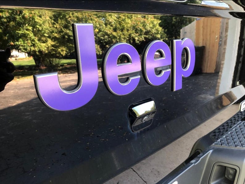 May include: Close-up of a black vehicle's rear with a purple and silver "Jeep" emblem. The emblem is three-dimensional and has a metallic finish. The background shows a blurred view of trees and a building.
