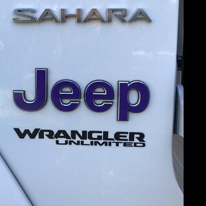 May include: Close-up of a white vehicle panel with the word "SAHARA" in silver lettering at the top. Below, the word "Jeep" is in purple with a silver outline, and "WRANGLER UNLIMITED" is in black.