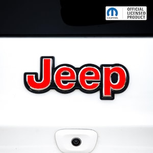 May include: A red and silver Jeep emblem with a black border is affixed to a white surface. The emblem is a licensed product, as indicated by the "Official Licensed Product" text and the Mopar logo in the upper right corner.