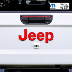 May include: White vehicle tailgate with a red "Jeep" emblem. The tailgate features a black handle, a red light, and a backup camera. The upper right corner displays the "Mopar" logo and "Official Licensed Product" text.