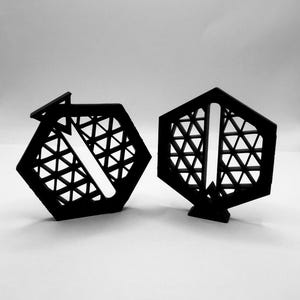 May include: Two black hexagonal objects with an intricate internal lattice structure. Each piece features a solid outer frame and a central vertical slot. The design consists of repeating triangular patterns within the hexagon, creating a geometric grid.