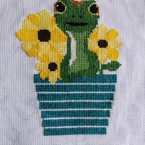 May include: Cross-stitch embroidery of a green frog with brown eyes sitting in a blue pot with yellow sunflowers.