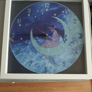 May include: Square white-framed wall clock with a celestial design. The clock face features a woman seated on a crescent moon, surrounded by stars against a blue background. The clock hands are red and black.