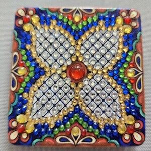 May include: A square-shaped mosaic art piece with a central red jewel surrounded by a white diamond pattern. The border is decorated with colorful beads in shades of blue, green, yellow, and red.
