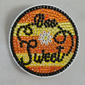 May include: A round, black and white sticker with a yellow and orange background. The sticker says "Make it Sweet" with a white daisy in the center.