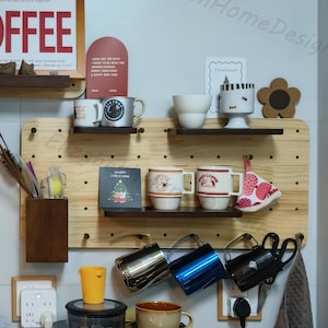 Op de afbeelding: Een houten pegboard met twee planken, versierd met mokken, een kerstkaart en een klein bloemetje. Er hangen ook verschillende metalen kannen aan haken aan het pegboard. Het pegboard is gemonteerd op een witte muur.