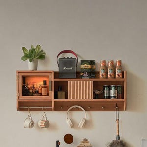 Può includere: Un ripiano a muro in legno con un armadio, ripiani aperti e ganci. Il ripiano è decorato con un piccolo altoparlante nero, capsule di caffè, bottiglie e un paio di cuffie. Il ripiano è montato su una parete bianca.