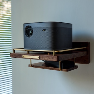 May include: A dark gray projector sits on a wooden shelf with a brass frame. The shelf is mounted to a white wall.
