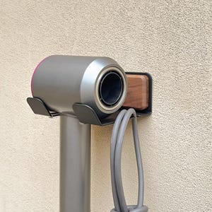 May include: A gray and pink hair dryer mounted on a wall with a black metal holder. The hair dryer is cylindrical with a circular nozzle. A gray cord hangs below the dryer. A wooden block is mounted next to the dryer.