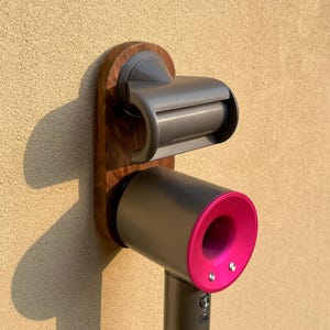 May include: A wooden wall-mounted holder for a hair dryer. The holder has a dark wood finish and holds a gray and pink hair dryer. The holder also has a built-in shelf for a roll of paper towels.