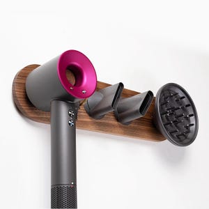 May include: A wall-mounted hair dryer holder made of dark wood with a grey hair dryer and three attachments. The hair dryer has a pink nozzle.