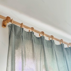 Puede incluir: Una barra de cortina de madera con anillos de madera clara y una cortina gris clara colgando de ella.
