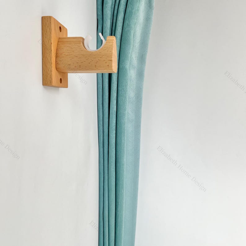 May include: A wooden curtain rod bracket mounted on a white wall with a teal curtain hanging to the right. The bracket is made of natural wood and has a simple design.