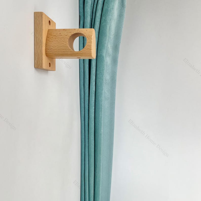 May include: A wooden curtain rod bracket with a round opening for the rod. The bracket is mounted on a white wall and a teal curtain is hanging to the right of the bracket.