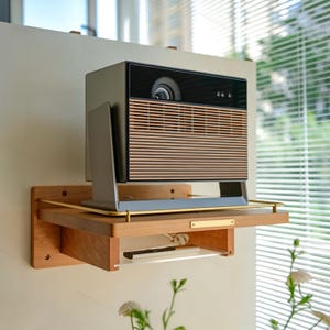 May include: A rectangular digital projector with a dark gray frame and a horizontal slatted wood-tone front panel, mounted on a light-colored wooden wall shelf featuring a small brass nameplate and a lower clear acrylic storage ledge.