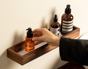 May include: A wooden shelf with a silver metal grid insert, holding three bottles of toiletries. The bottles are clear glass with brown liquid and black pump tops. The shelf is mounted on a white wall.