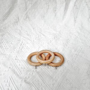 May include: Three wooden curtain rings with a natural finish. The rings are stacked on top of each other and have a silver hook on each side.