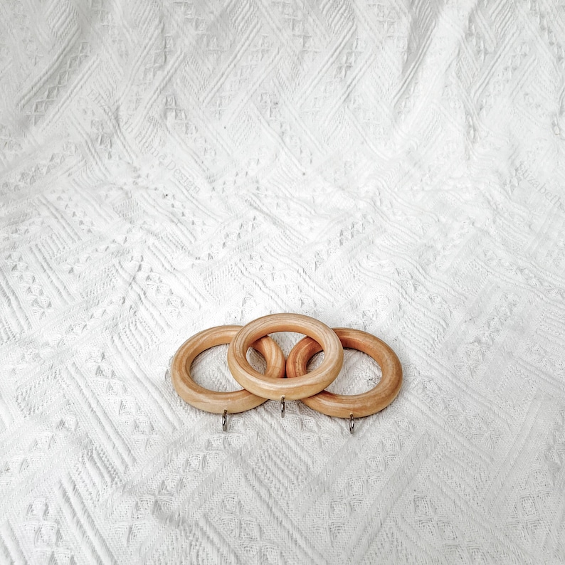 May include: Three wooden curtain rings with a natural finish. The rings are stacked on top of each other and have a silver hook on each side.