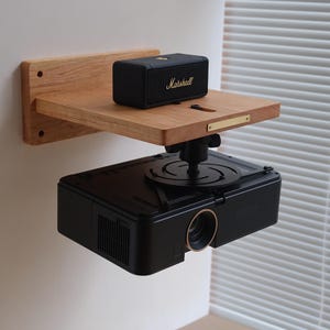 Op de afbeelding: Een houten wandplank met een zwarte speaker met het label "Marshall" erop en een zwarte projector die eronder is gemonteerd. De plank is bevestigd aan een witte muur.
