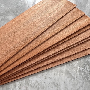 May include: A stack of thin, rectangular wooden planks with a reddish-brown hue. The wood grain is visible, and the planks are arranged in a fan-like pattern. The background is a mottled gray surface.