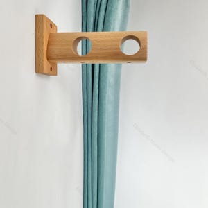May include: A wooden curtain rod bracket with three holes. The bracket is mounted on a white wall and a light blue curtain is hanging from it.