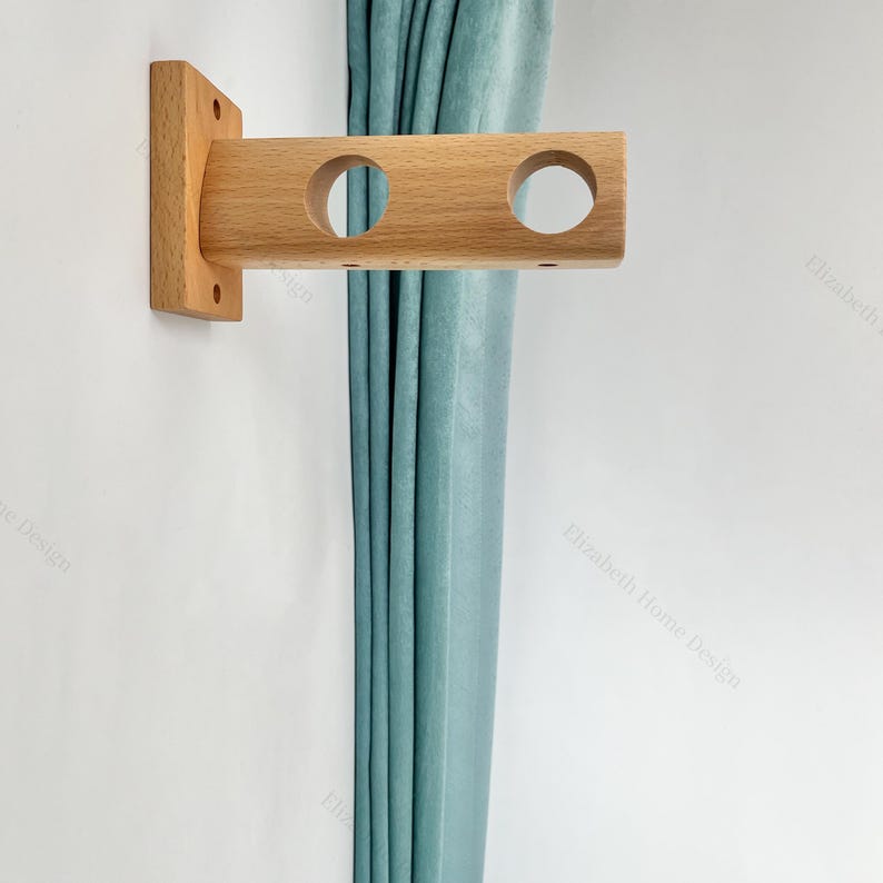 May include: A wooden curtain rod bracket with three holes. The bracket is mounted on a white wall and a light blue curtain is hanging from it.