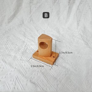 May include: Wooden wall mount with a round opening, measuring 8.9 cm by 9.5 cm. The mount is made of light brown wood and has a simple design.