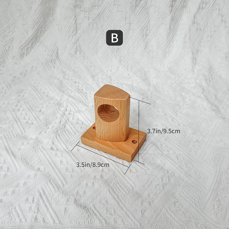 May include: Wooden wall mount with a round opening, measuring 8.9 cm by 9.5 cm. The mount is made of light brown wood and has a simple design.