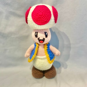 May include: A crocheted plush toy of a mushroom character with a red and white hat, blue vest, and brown shoes. The character has a smiling face with black eyes and a red mouth.