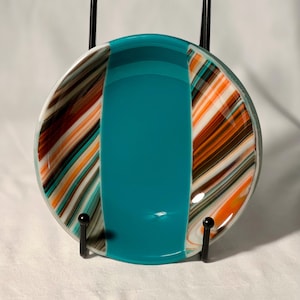 May include: A circular fused glass dish featuring a wide, solid teal center panel flanked by two side sections with diagonal, swirling stripes of orange, brown, white, and teal. The dish is held upright by a black metal wire display stand.
