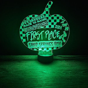 May include: Green illuminated trophy shaped like a checkered flag with the text "1st Annual Race to Raise Swapathon 2024 First Place Sand Springs BMX"