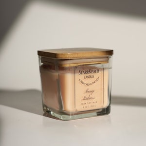 May include: A square glass candle jar with a wooden lid. The candle is peach-colored and labeled "StarrKissed Candles Mango Madness." The candle is 8.5 oz (240g) and made of 100% soy wax.
