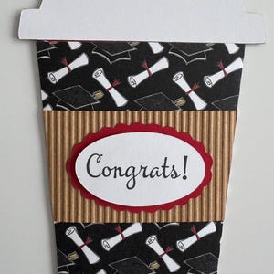 May include: A graduation-themed greeting card shaped like a coffee cup. The card features a white lid, a black background with graduation caps and scrolls, a corrugated brown sleeve, and a red-edged oval with the word "Congrats!"
