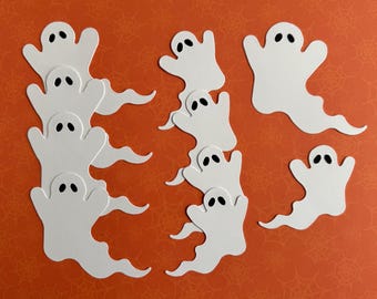 May include: White ghost cutouts with black oval eyes on an orange background with a spiderweb pattern. The ghosts have a simple design, suitable for Halloween decorations or crafts. Several ghost shapes are visible in the image.