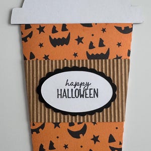 May include: A Halloween-themed paper coffee cup design. The cup features an orange background with black jack-o'-lantern faces and stars. The cup has a brown corrugated sleeve and a white lid. The words "happy HALLOWEEN" are printed on an oval label.