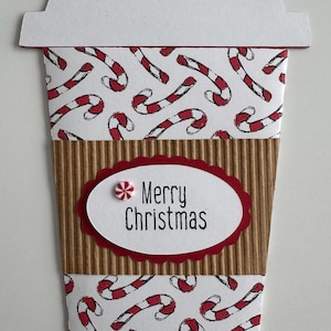 May include: A Christmas-themed paper coffee cup design. The cup features a white lid, a corrugated cardboard midsection, and a candy cane pattern. The words "Merry Christmas" are displayed on a red-edged oval.