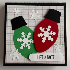 May include: A handmade Christmas card featuring a red and green light bulb design with white snowflake accents. The card includes the text "JUST A NOTE" on a white rectangular label. The background has a grid pattern.